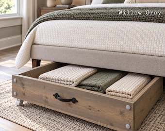 May include: A rectangular under-bed storage drawer made of light brown wood with a visible grain. The drawer features a black metal handle, four white caster wheels, and four small circular corner bumpers. It contains three folded blankets.
