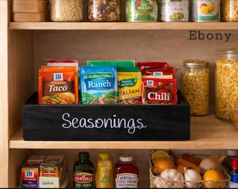 May include: A black wooden storage bin with the word Seasonings written in white script on the front. The bin is filled with various colorful seasoning packets, including taco, ranch, and chili flavors, organized on a wooden pantry shelf.