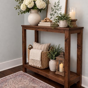 May include: A dark walnut console table with a rectangular top and a lower shelf. The table holds a white vase with white flowers, a framed leaf print, a wooden chain link, a small potted plant, and a candle in a glass holder on the top shelf.