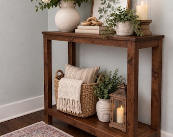 May include: A dark walnut console table with a rectangular top and a lower shelf. The table holds a white vase with white flowers, a framed leaf print, a wooden chain link, a small potted plant, and a candle in a glass holder on the top shelf.