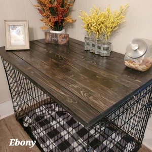 May include: A dark stained wooden dog crate with a black wire frame. The crate has a black and white plaid blanket inside. The top of the crate is decorated with fall-themed floral arrangements, a framed photo, and a jar of dog treats. The word "Ebony" is visible.