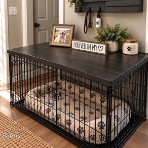 Pu&ograve; includere: Una gabbia per cani in metallo nero con un piano superiore in legno scuro. All'interno si trova una cuccia beige con un motivo a impronte nere. Sul piano in legno poggiano una cornice, un cartello con scritto "FOREVER IN MY ♡", una pianta e un barattolo.