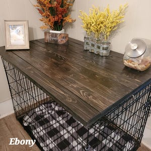 Pu&ograve; includere: Una gabbia per cani in filo metallico nero sormontata da una tavola di legno tinta di marrone scuro. All'interno c'&egrave; una coperta a quadri bianchi e neri. La superficie superiore ospita una cornice, un cartello My Farmhouse Essentials e un barattolo.