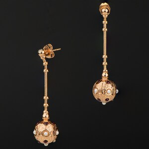 May include: A pair of gold dangle earrings with a round, textured ball at the end of each earring. The balls are adorned with small white pearls and red gemstones.