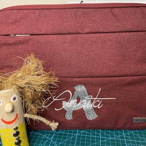 Puede incluir: Funda para portátil de color burdeos con una letra A de purpurina plateada y el nombre "Anita" escrito en cursiva. La funda tiene un bolsillo con cremallera y un pequeño logotipo que dice "Yooya".