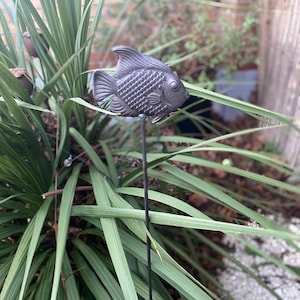 Puede incluir: Una estaca de jardín de metal negro con un diseño de pez. El pez está detallado con escamas y tiene un aspecto realista. La estaca está insertada en un lecho de plantas verdes.