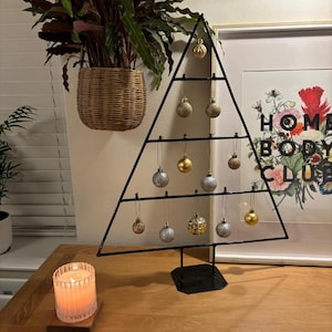 May include: A black metal Christmas tree stand with silver and gold baubles hanging from it. The stand is on a wooden table with a lit candle in front of it.