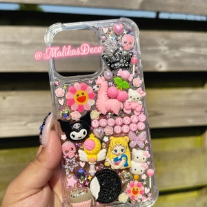 Può includere: Una custodia per telefono trasparente decorata con abbellimenti rosa, bianchi e neri. La custodia presenta una varietà di ciondoli, tra cui un lama rosa, un fiore, un teschio, una corona, un personaggio Hello Kitty, un personaggio Sailor Moon e un biscotto bianco e nero. La custodia include anche il testo "MALKAS DECO" in lettere rosa.