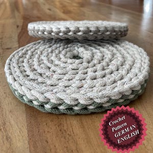 May include: Two crocheted coasters, one on top of the other. The top coaster is white with a green border. The bottom coaster is gray with a white border. The coasters are on a wooden surface. The text "Crochet Pattern GERMAN ENGLISH" is on a red starburst sticker.