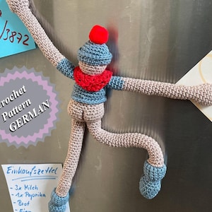 May include: A crocheted doll with a blue hat and red scarf is attached to a silver refrigerator with magnets. The doll has long arms and legs. There are several notes on the refrigerator, including one that says "Crochet Pattern GERMAN" and another that says "Oma anrufen!"