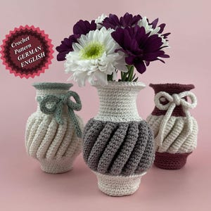 Peut inclure: Trois vases en crochet de différentes couleurs, avec un bouquet de fleurs blanches et violettes dans le vase du milieu. Les vases sont tous dans un style similaire, avec un corps rond et un sommet évasé. Le vase du milieu est gris et blanc, le vase de gauche est blanc et le vase de droite est violet et blanc.