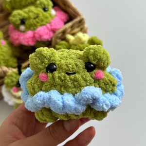 May include: A handmade green crochet frog with black eyes and pink cheeks. The frog has a blue and white ruffled trim. Other similar frogs are in a woven basket, some with pink accents.