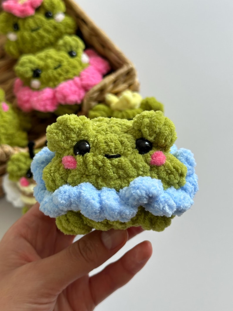 May include: A handmade green crochet frog with black eyes and pink cheeks. The frog has a blue and white ruffled trim. Other similar frogs are in a woven basket, some with pink accents.