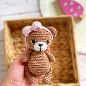 May include: A brown crocheted teddy bear wearing a pink bow. The bear is sitting in a wicker basket.