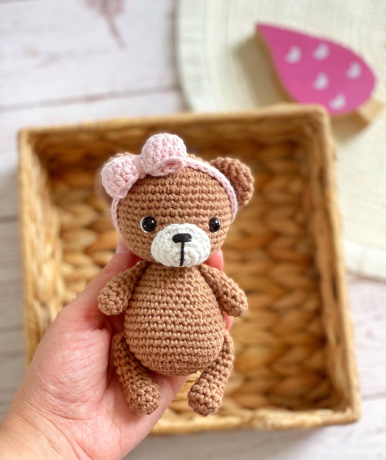 May include: A brown crocheted teddy bear wearing a pink bow. The bear is sitting in a wicker basket.