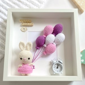 May include: A white frame with a crocheted bunny holding purple, pink, and white balloons. The frame also includes a small white clock and wooden blocks spelling "Julia Maria".