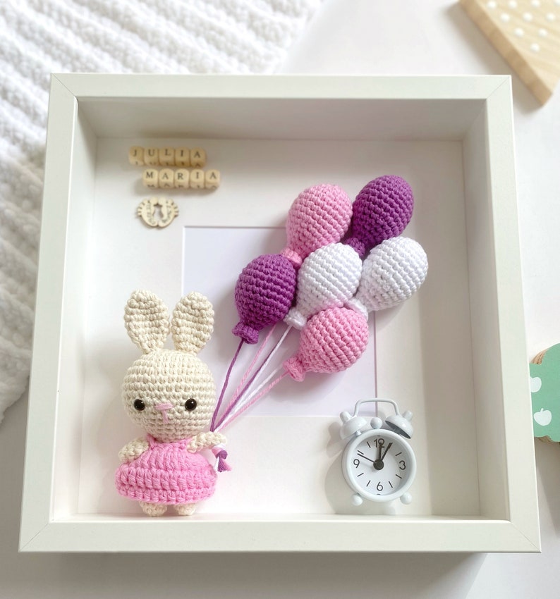 May include: A white frame with a crocheted bunny holding purple, pink, and white balloons. The frame also includes a small white clock and wooden blocks spelling "Julia Maria".