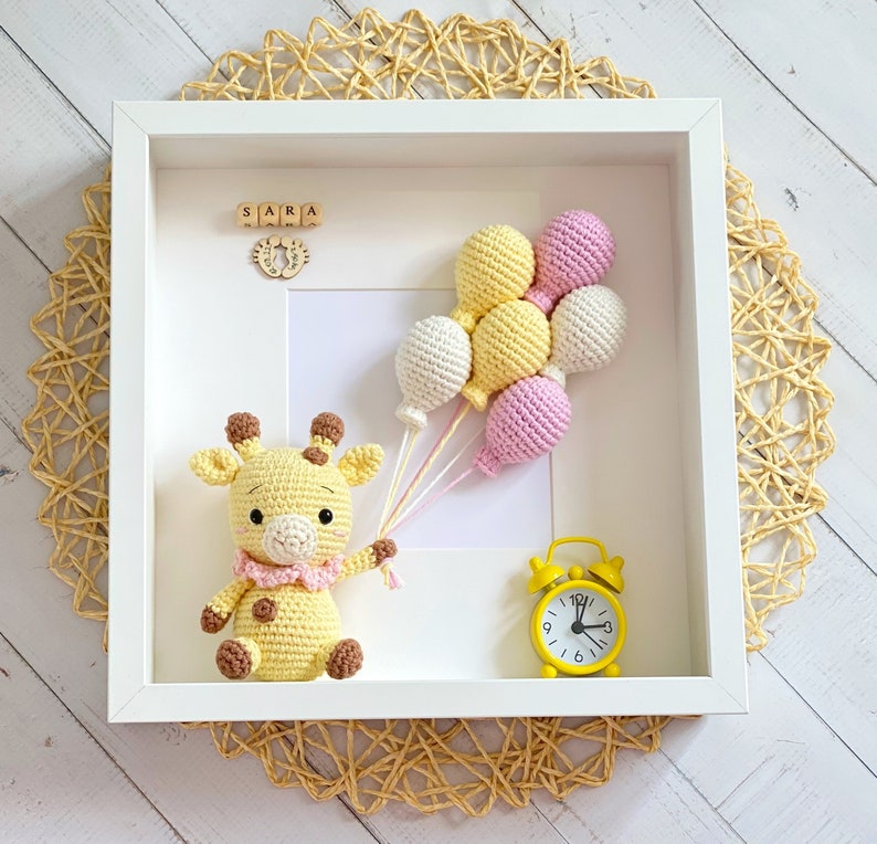 May include: A white frame with a yellow and pink crocheted giraffe toy, a yellow alarm clock, and a cluster of yellow and pink crocheted balloons. The frame has the word "SARA" spelled out in wooden blocks.