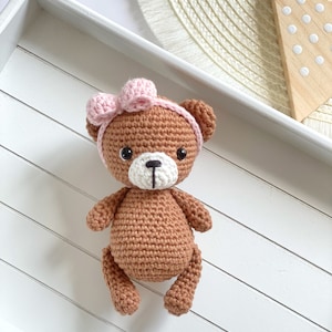 May include: A brown crocheted teddy bear with a pink bow on its head. The bear is sitting on a white wooden tray.