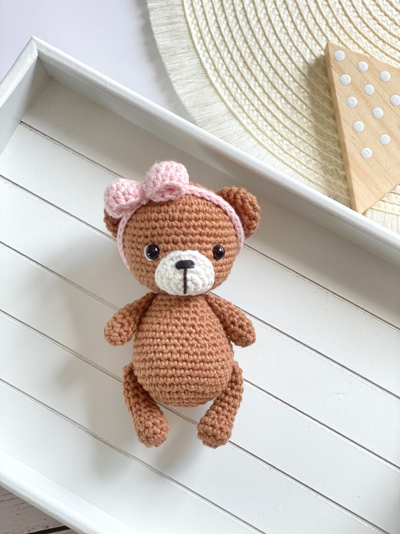 May include: A brown crocheted teddy bear with a pink bow on its head. The bear is sitting on a white wooden tray.
