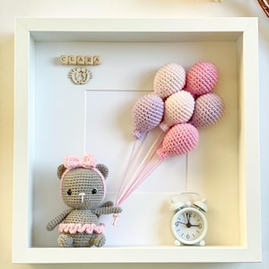 May include: A white frame with a gray crocheted teddy bear holding pink and purple balloons. The frame also includes a wooden block name sign that reads "CLARA" and a small white alarm clock.