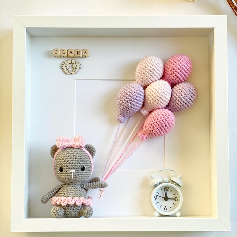 May include: A white frame with a gray crocheted teddy bear holding pink and purple balloons. The frame also includes a wooden block name sign that reads "CLARA" and a small white alarm clock.