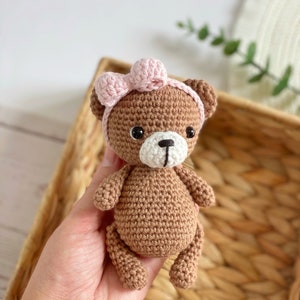 May include: A brown crocheted teddy bear wearing a pink bow. The bear has black eyes and a white nose and mouth.