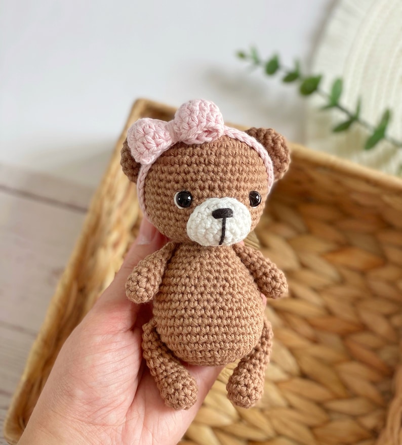 May include: A brown crocheted teddy bear wearing a pink bow. The bear has black eyes and a white nose and mouth.