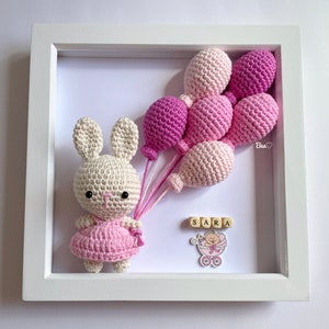 May include: A white crocheted bunny holding pink balloons in a white frame. The frame also includes a pink baby carriage with a baby inside and the name "SARA" spelled out in wooden blocks.