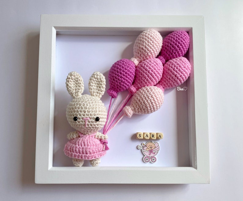 May include: A white crocheted bunny holding pink balloons in a white frame. The frame also includes a pink baby carriage with a baby inside and the name "SARA" spelled out in wooden blocks.