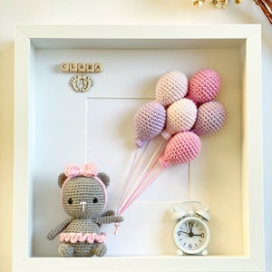 May include: A white frame with a crocheted teddy bear holding pink and purple balloons. The frame also includes a wooden name block that reads "CLARA" and a small white alarm clock.