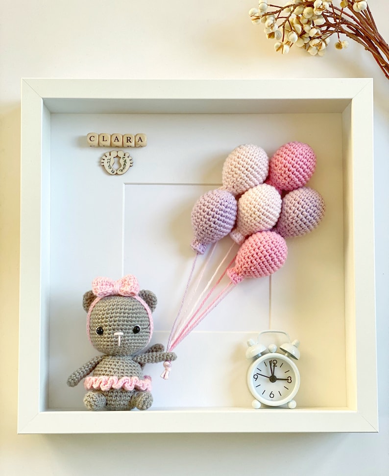 May include: A white frame with a crocheted teddy bear holding pink and purple balloons. The frame also includes a wooden name block that reads "CLARA" and a small white alarm clock.