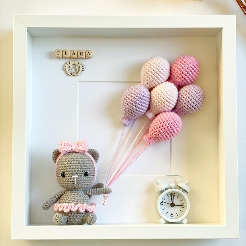 May include: A white frame with a gray crocheted teddy bear holding pink and purple balloons. The frame also includes a wooden block name sign that reads "CLARA" and a small white alarm clock.