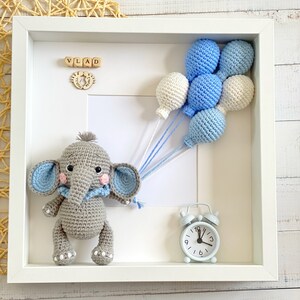 May include: A white picture frame with a gray crocheted elephant holding blue and white balloons. The frame also includes a small blue alarm clock and wooden blocks spelling "VLAD".