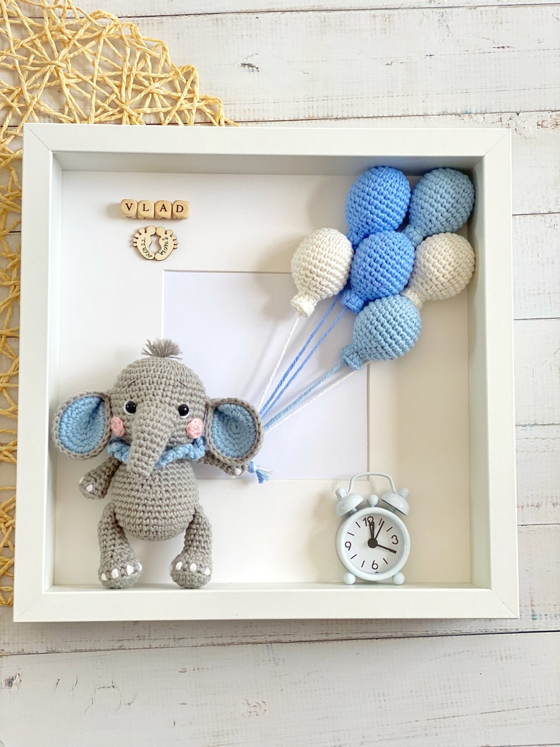 May include: A white picture frame with a gray crocheted elephant holding blue and white balloons. The frame also includes a small blue alarm clock and wooden blocks spelling "VLAD".