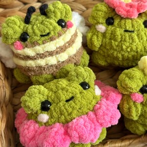 May include: Close-up of several handmade crochet plush toys. The toys are green with black eyes and pink cheeks. One is designed as a bee with brown and yellow stripes, and another has a pink flower on its head. The toys are in a woven basket.