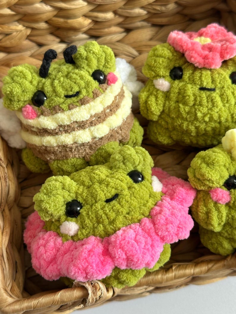 May include: Close-up of several handmade crochet plush toys. The toys are green with black eyes and pink cheeks. One is designed as a bee with brown and yellow stripes, and another has a pink flower on its head. The toys are in a woven basket.
