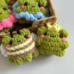 May include: A collection of handmade green crochet frog plushies, some with accessories. One frog has bee-like stripes, another has a blue ruffle, and another has a yellow crown. The frogs have black eyes and pink cheeks, and are in a woven basket.