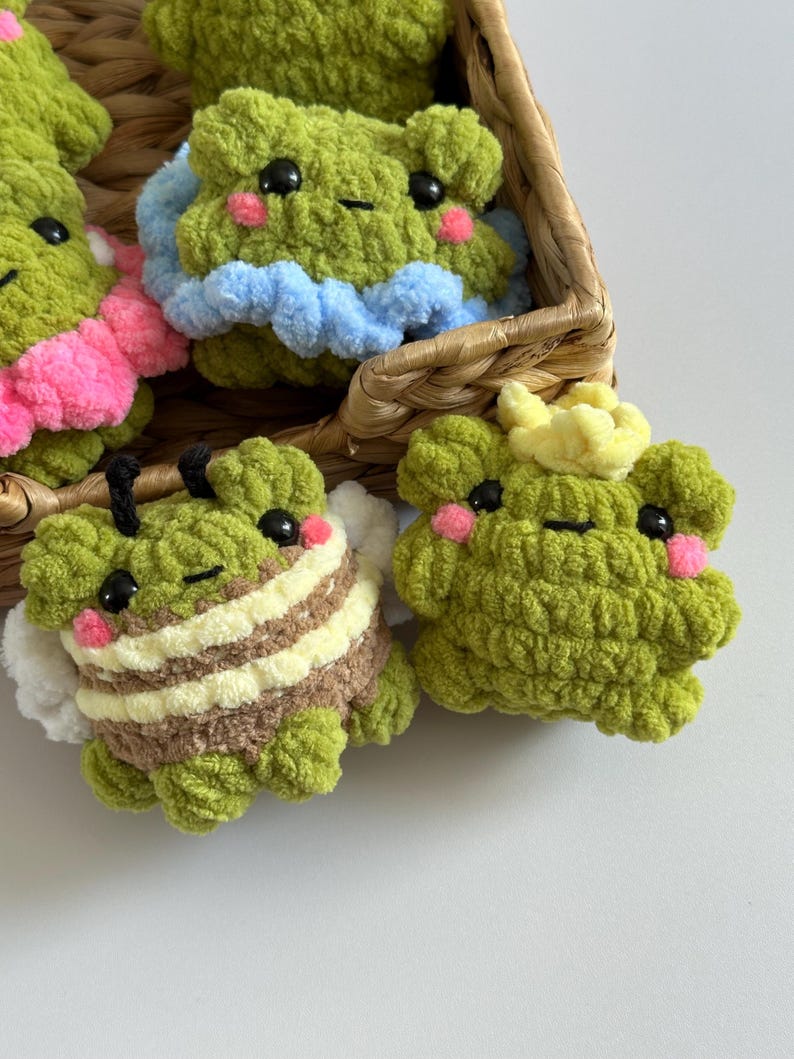 May include: A collection of handmade green crochet frog plushies, some with accessories. One frog has bee-like stripes, another has a blue ruffle, and another has a yellow crown. The frogs have black eyes and pink cheeks, and are in a woven basket.