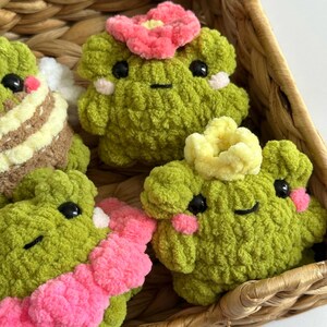 May include: A collection of handmade green crochet frog plushies with black eyes and pink blush. Some frogs have pink or yellow flower accents. The frogs are displayed in a woven basket.