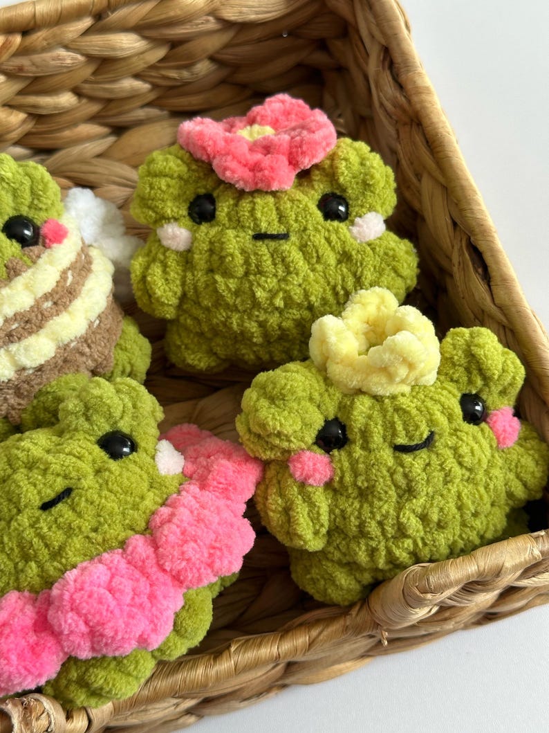 May include: A collection of handmade green crochet frog plushies with black eyes and pink blush. Some frogs have pink or yellow flower accents. The frogs are displayed in a woven basket.