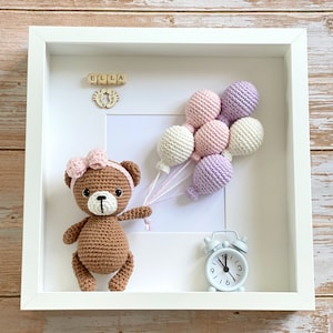 May include: A white picture frame with a brown crocheted teddy bear holding pink and purple balloons. The frame also includes a wooden name block that says "ELLA" and a small white alarm clock.