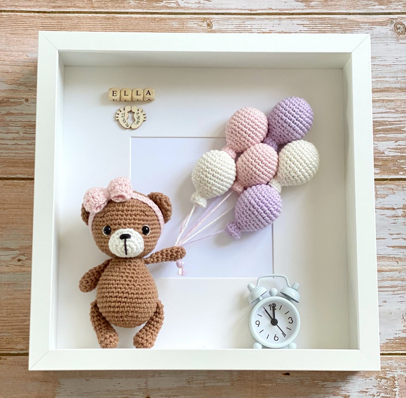 May include: A white picture frame with a brown crocheted teddy bear holding pink and purple balloons. The frame also includes a wooden name block that says "ELLA" and a small white alarm clock.
