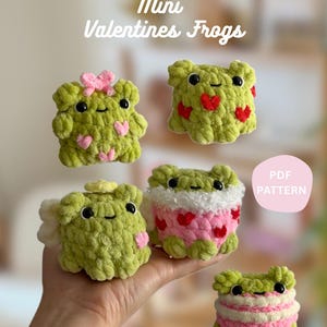May include: Five handmade, green crochet frogs with black eyes and smiling faces. Some frogs have pink heart accents, bows, or a white and pink striped design. The text "Mini Valentines Frogs" is at the top.