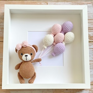 May include: A brown crocheted teddy bear holding a bunch of pastel pink, purple, and white crocheted balloons in a white picture frame.