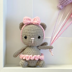 May include: A crocheted gray bear wearing a pink bow and a pink tutu holding a pink and white string attached to a purple balloon.