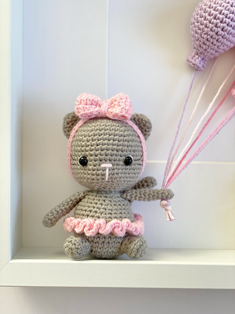 May include: A crocheted gray bear wearing a pink bow and a pink tutu holding a pink and white string attached to a purple balloon.