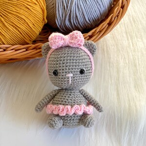 May include: A gray crocheted cat wearing a pink bow and a pink tutu. The cat is sitting on a white fluffy surface.