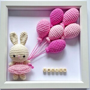 May include: A white crocheted bunny holding pink balloons with the name "AMALIA" spelled out in wooden blocks.