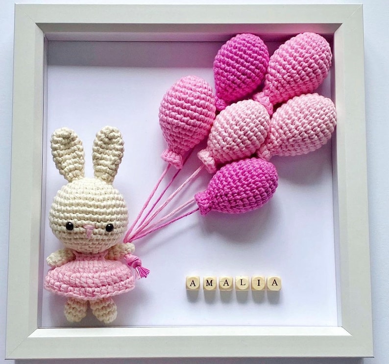 May include: A white crocheted bunny holding pink balloons with the name "AMALIA" spelled out in wooden blocks.